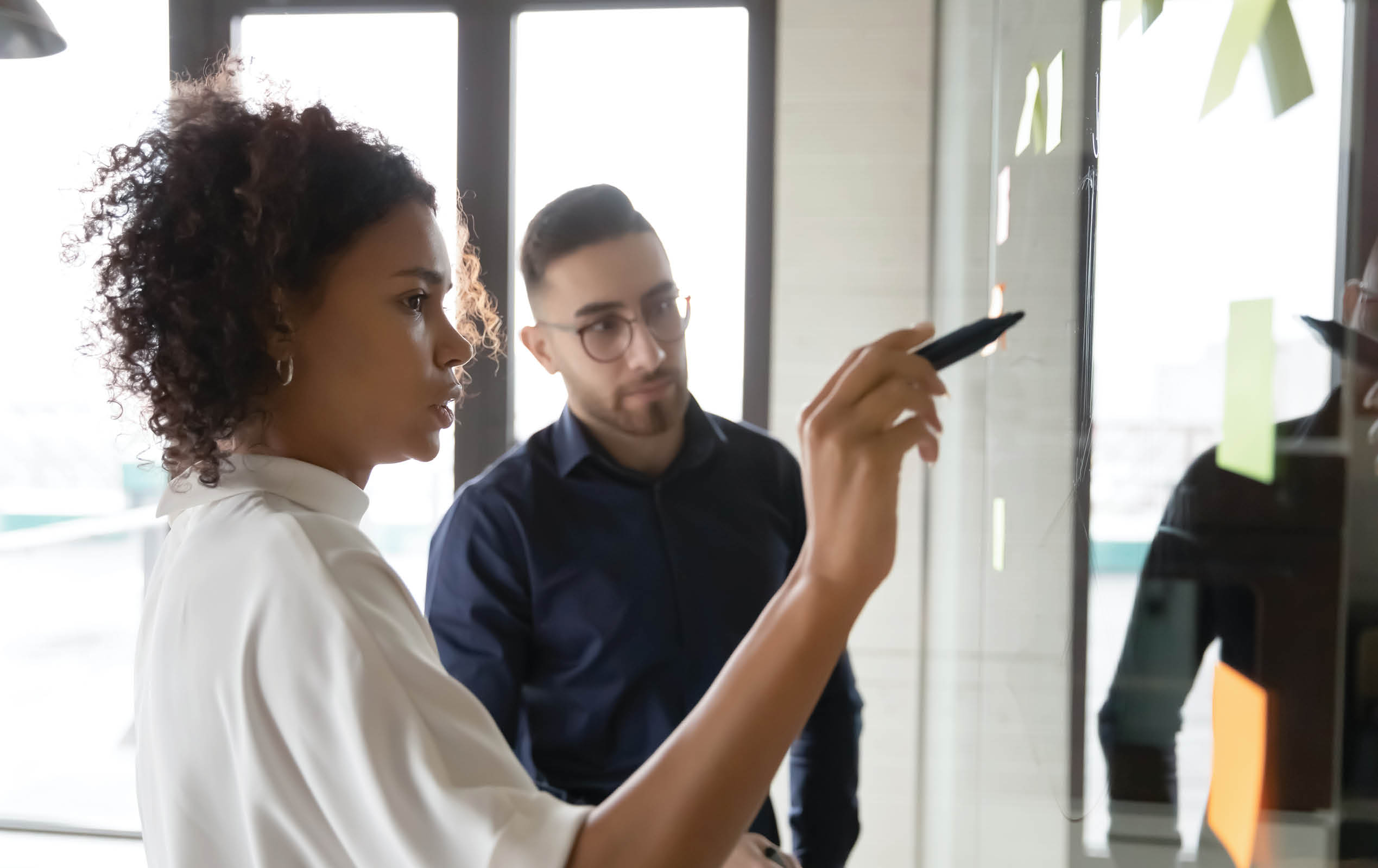 Focused multiracial employees write ideas on glass wall plan startup in office together, diverse colleagues engaged in creative thinking, develop project at meeting in boardroom, teamwork concept